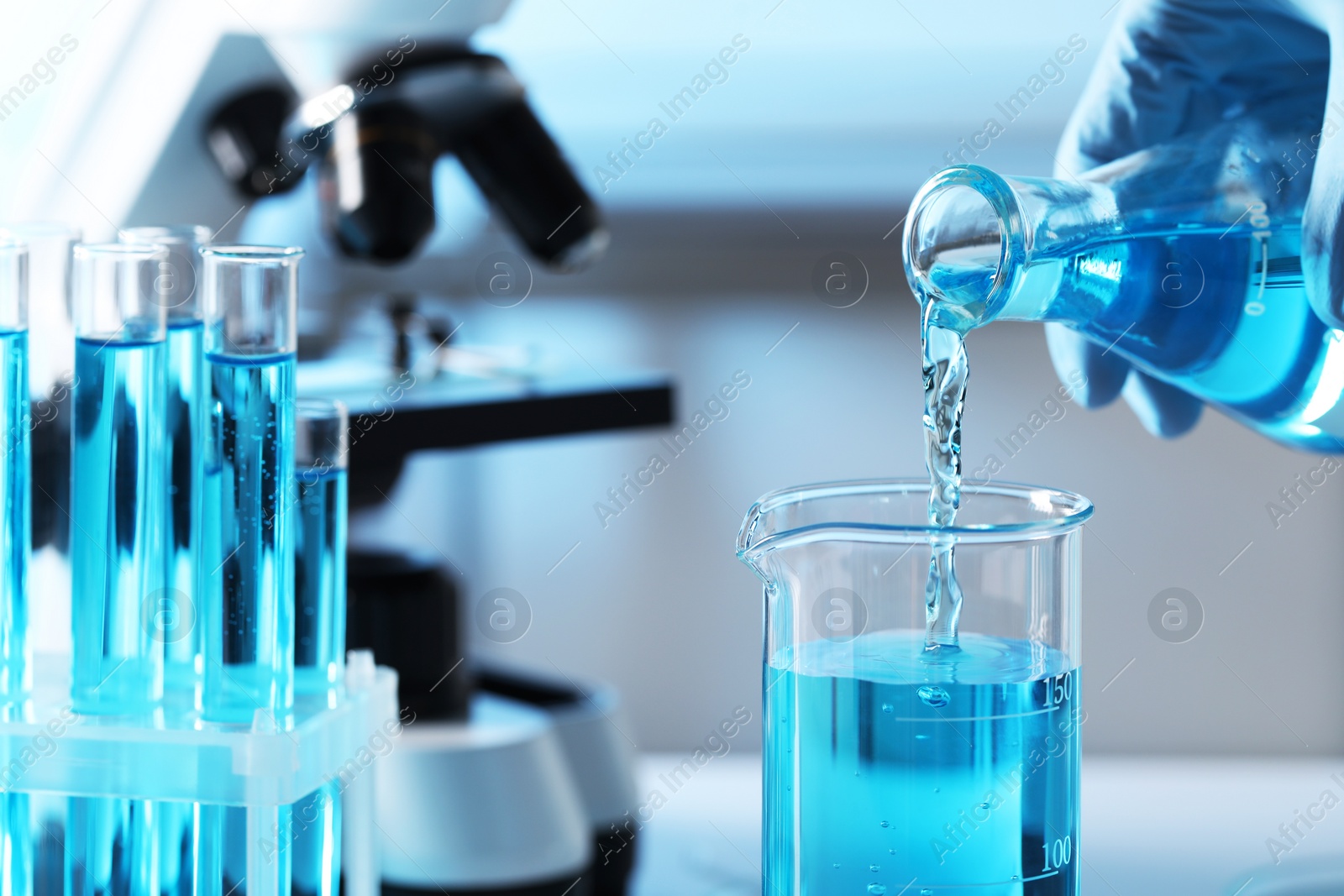 Photo of Scientist pouring light blue liquid from flask into beaker near test tubes in laboratory, closeup