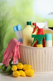 Photo of Spring cleaning. Plastic basket with detergents, supplies and beautiful flowers on white wooden table outdoors