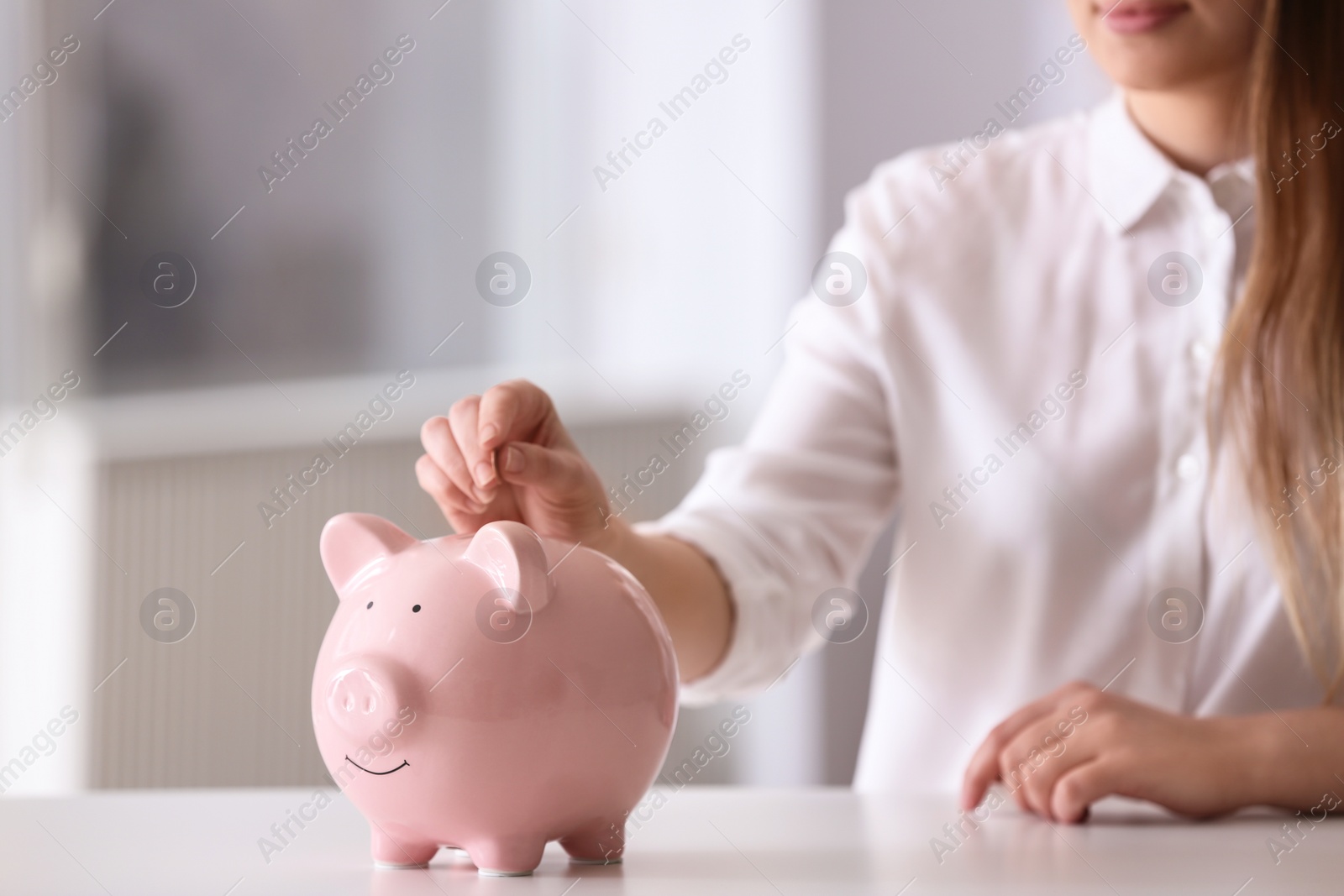 Photo of Woman putting coin into piggy bank at table indoors, closeup