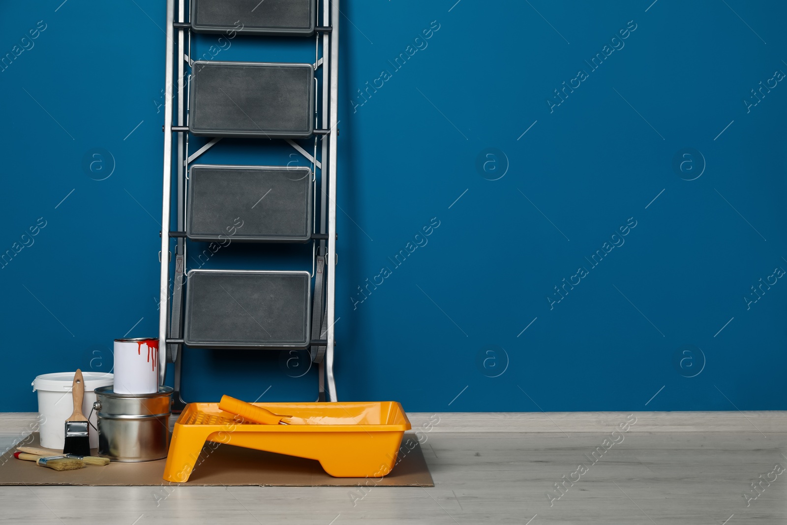 Photo of Metallic folding ladder and painting tools near blue wall indoors, space for text