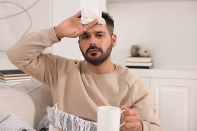 Photo of Sick man with tissue and cup of drink on sofa at home. Cold symptoms