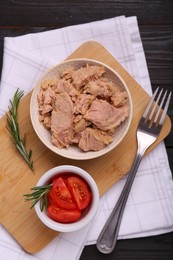 Photo of Bowl with canned tuna, tomatoes and rosemary on black wooden table, flat lay