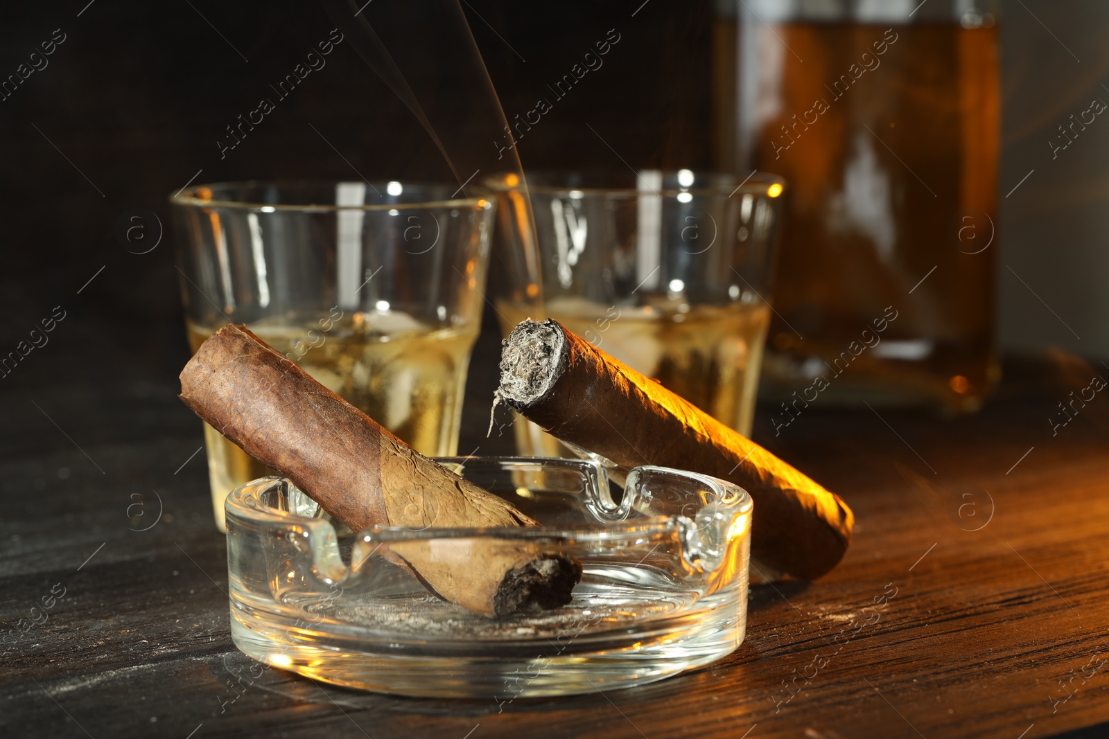 Photo of Cigars, ashtray and whiskey with ice cubes on black wooden table, closeup