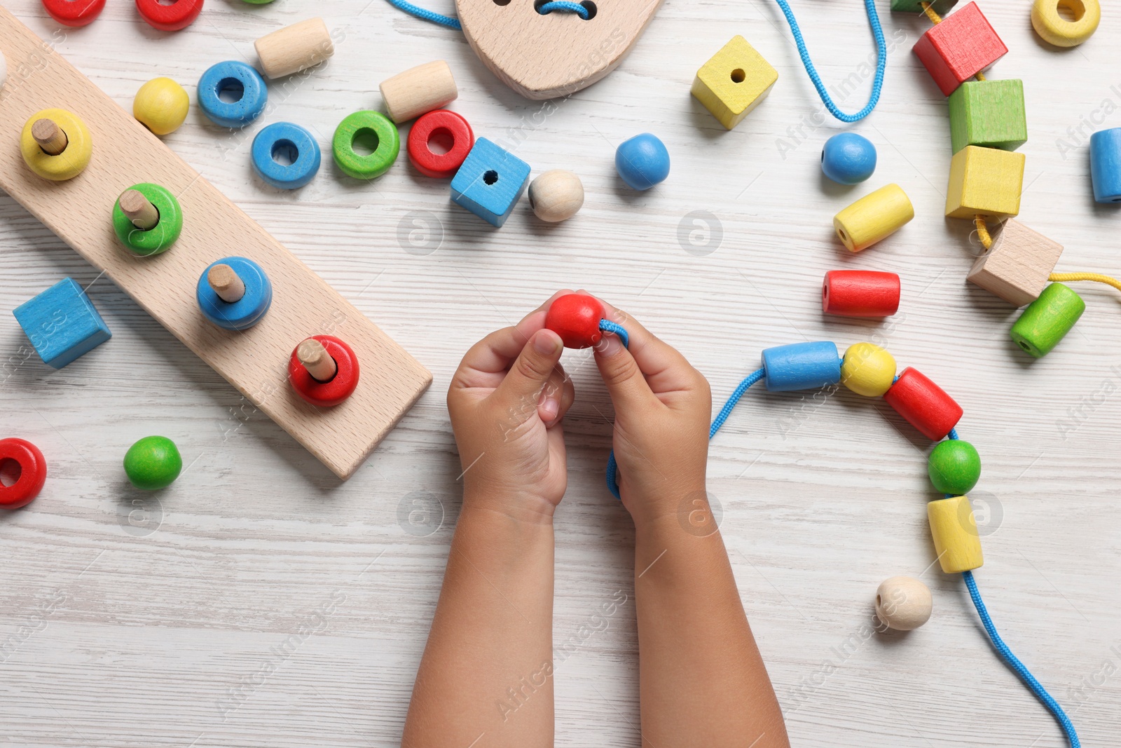 Photo of Motor skills development. Little child playing with wooden pieces and string for threading activity at white table, top view