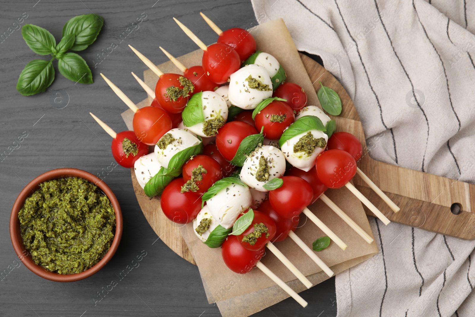 Photo of Caprese skewers with tomatoes, mozzarella balls, basil and pesto sauce on grey wooden table, flat lay