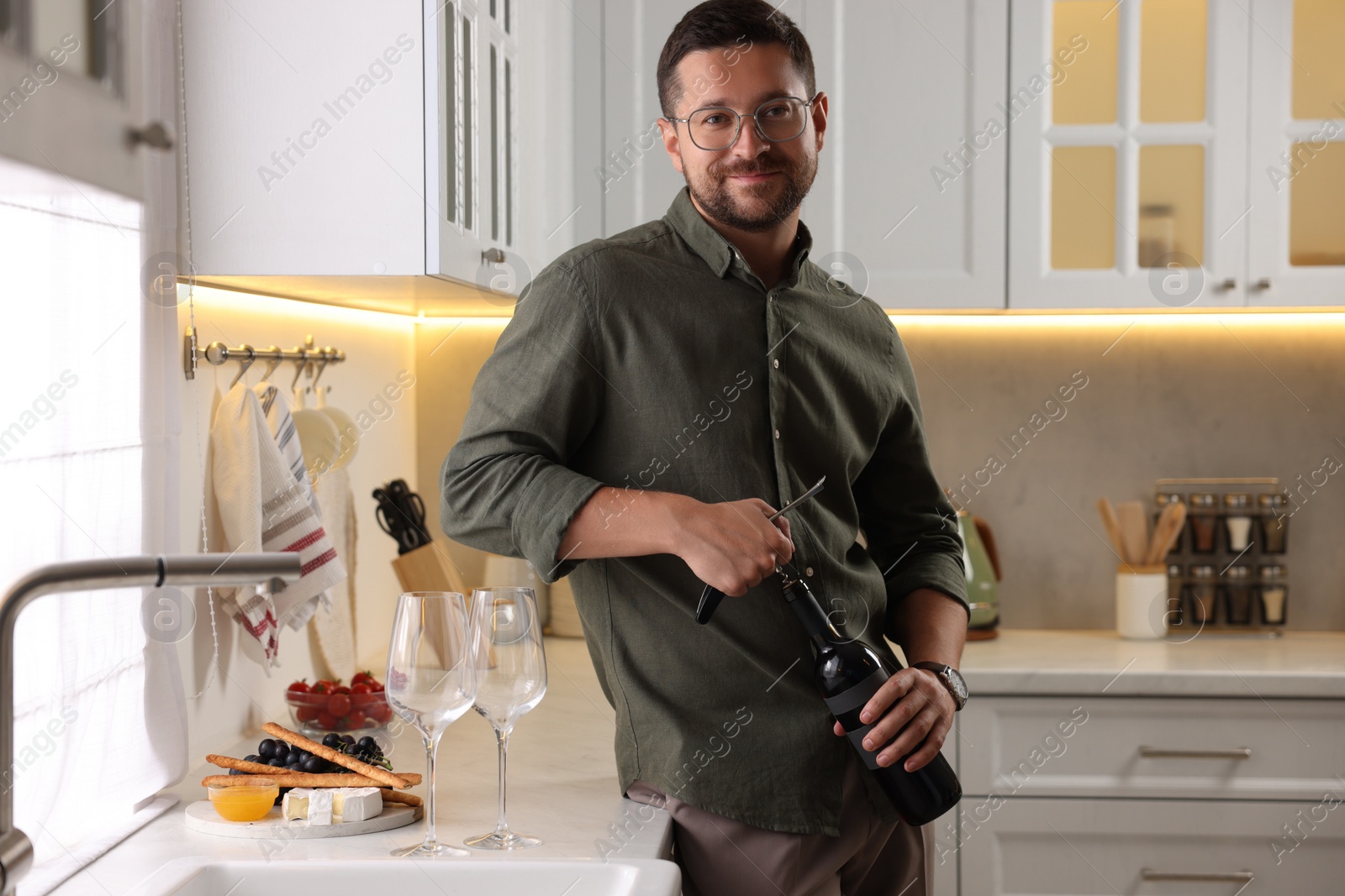 Photo of Romantic dinner. Man opening wine bottle with corkscrew in kitchen