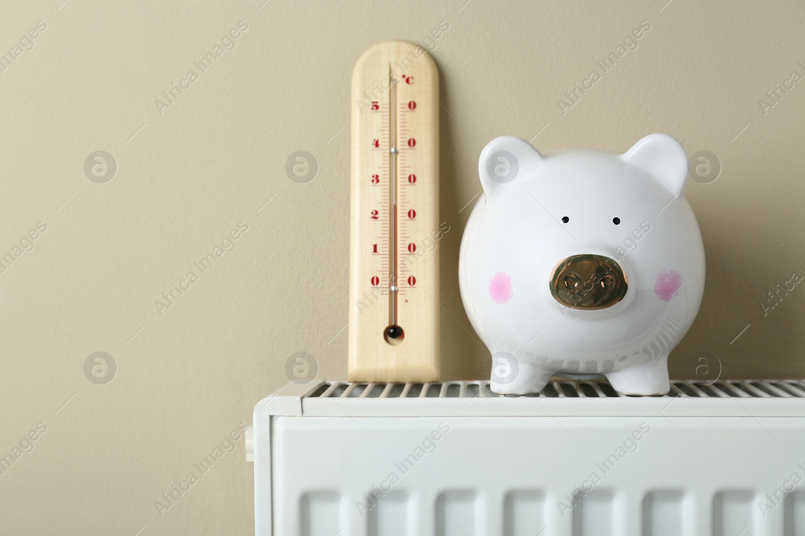 Photo of Piggy bank and thermometer on heating radiator against beige background