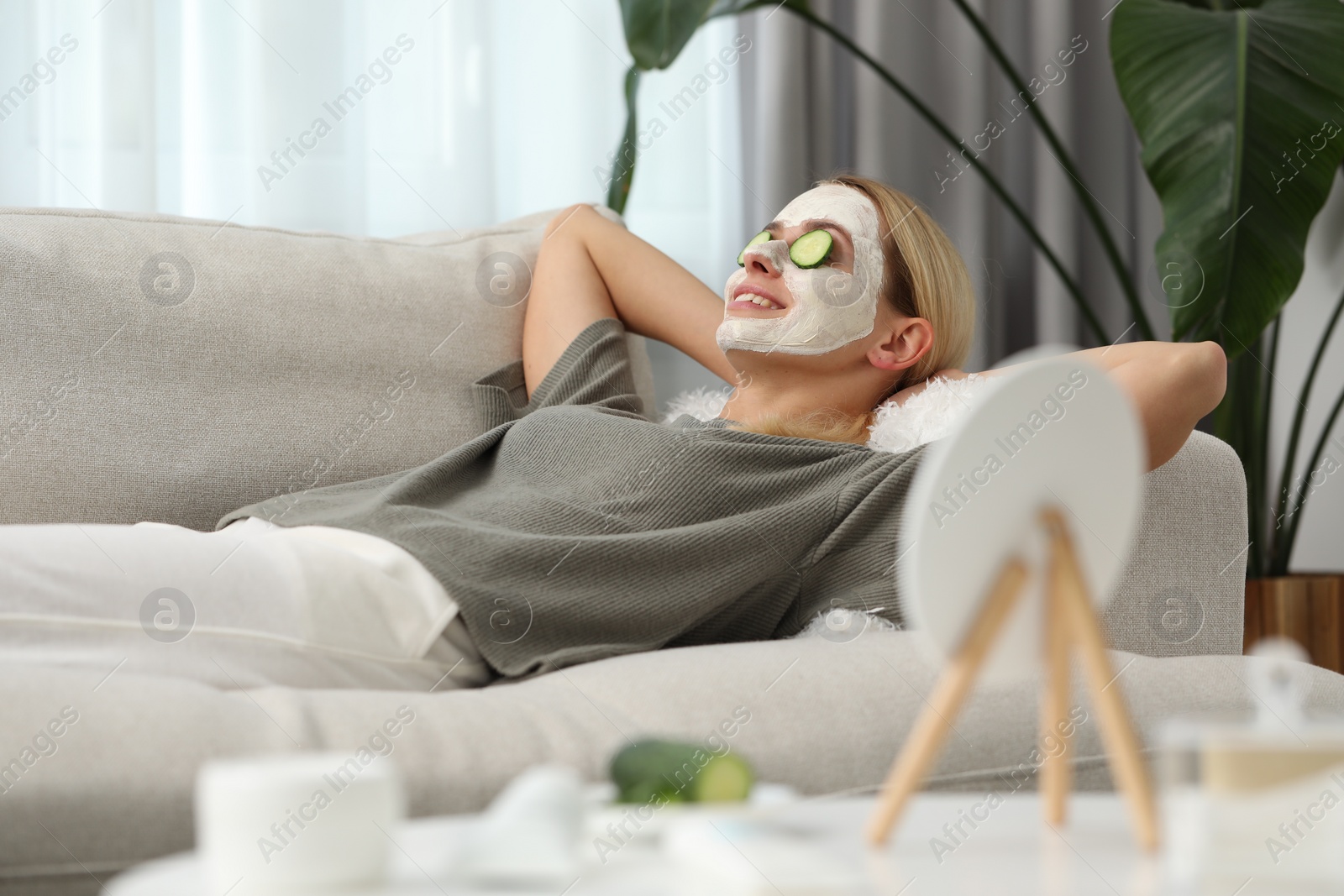 Photo of Young woman with face mask and cucumber slices resting on sofa at home. Spa treatments