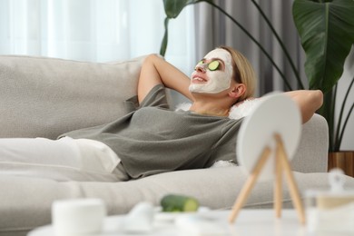 Photo of Young woman with face mask and cucumber slices resting on sofa at home. Spa treatments