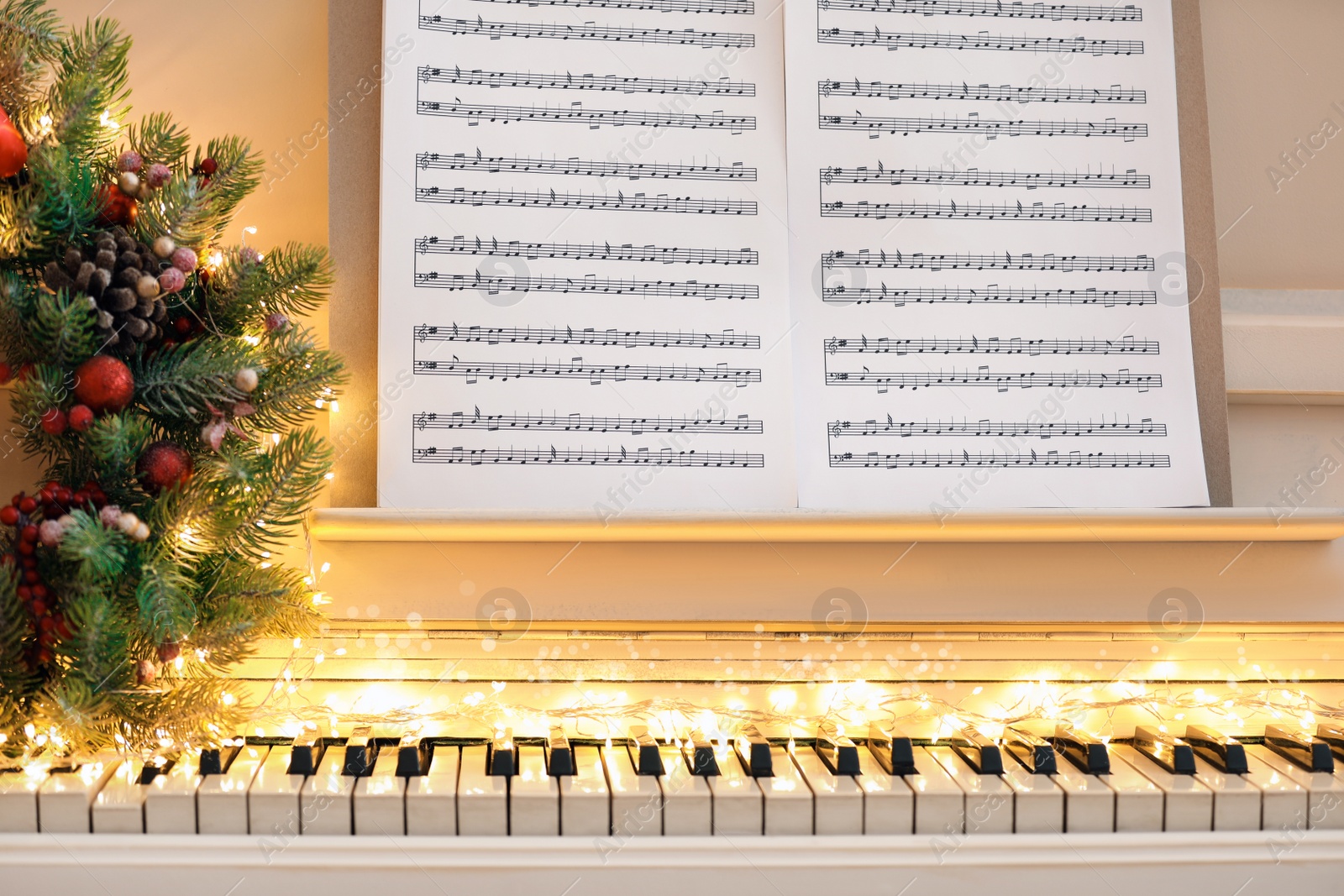 Photo of White piano with fairy lights, beautiful Christmas wreath and music sheets, closeup