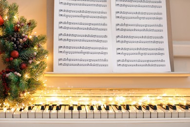 Photo of White piano with fairy lights, beautiful Christmas wreath and music sheets, closeup