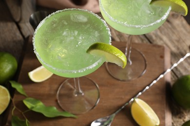 Delicious Margarita cocktail with ice cubes in glasses, limes and bar spoon on wooden table