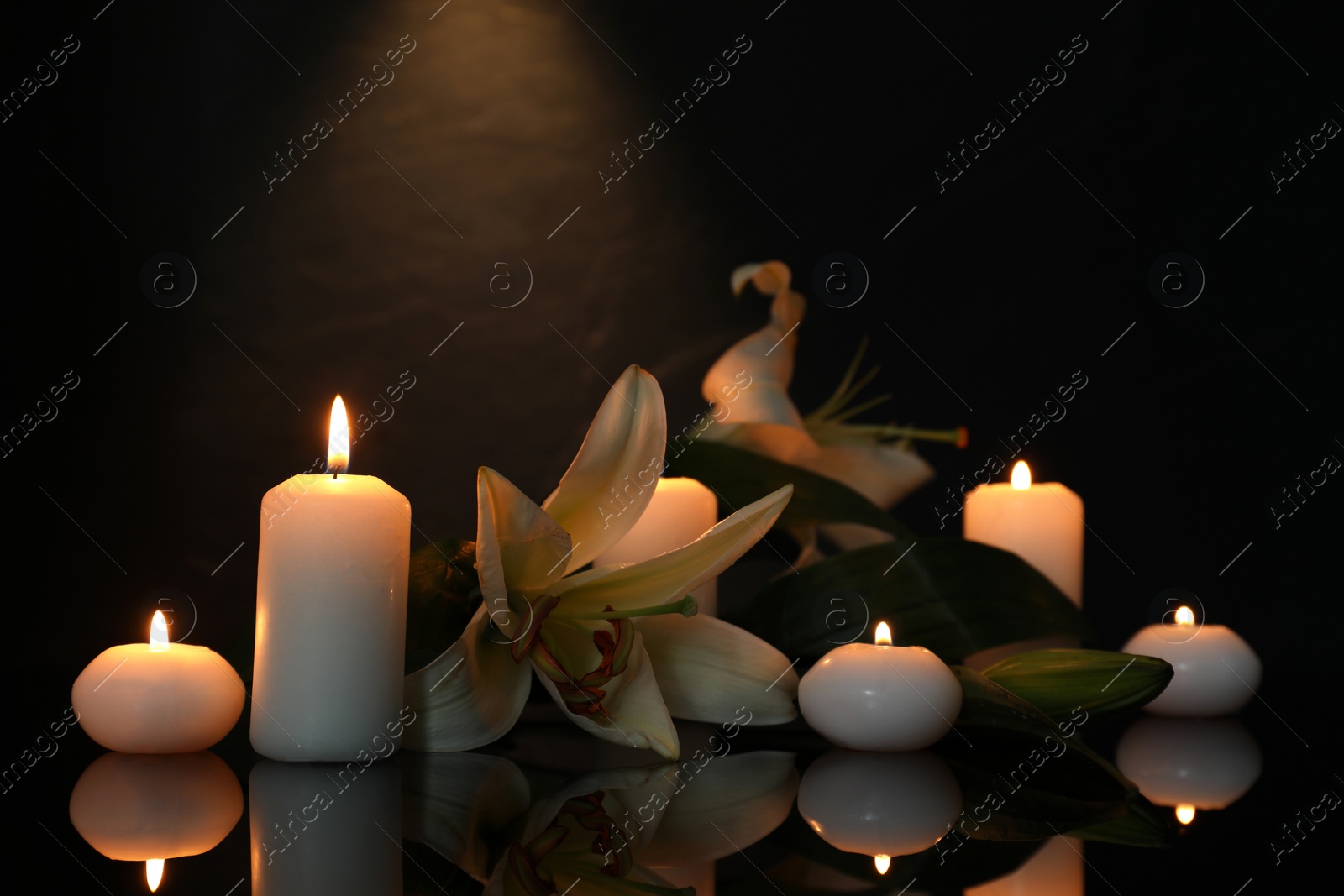 Photo of White lilies and burning candles on black mirror surface in darkness, space for text. Funeral symbols