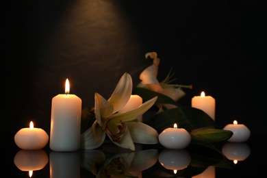 White lilies and burning candles on black mirror surface in darkness, space for text. Funeral symbols