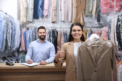 Dry-cleaning service. Happy woman holding hanger with coat in plastic bag and showing thumb up indoors. Worker taking notes at workplace