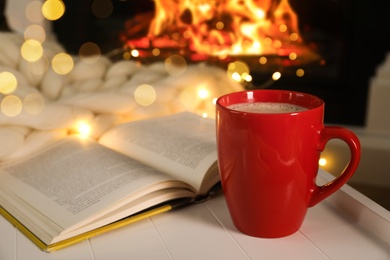 Photo of Cup of hot drink and book near fireplace. Cozy Christmas atmosphere