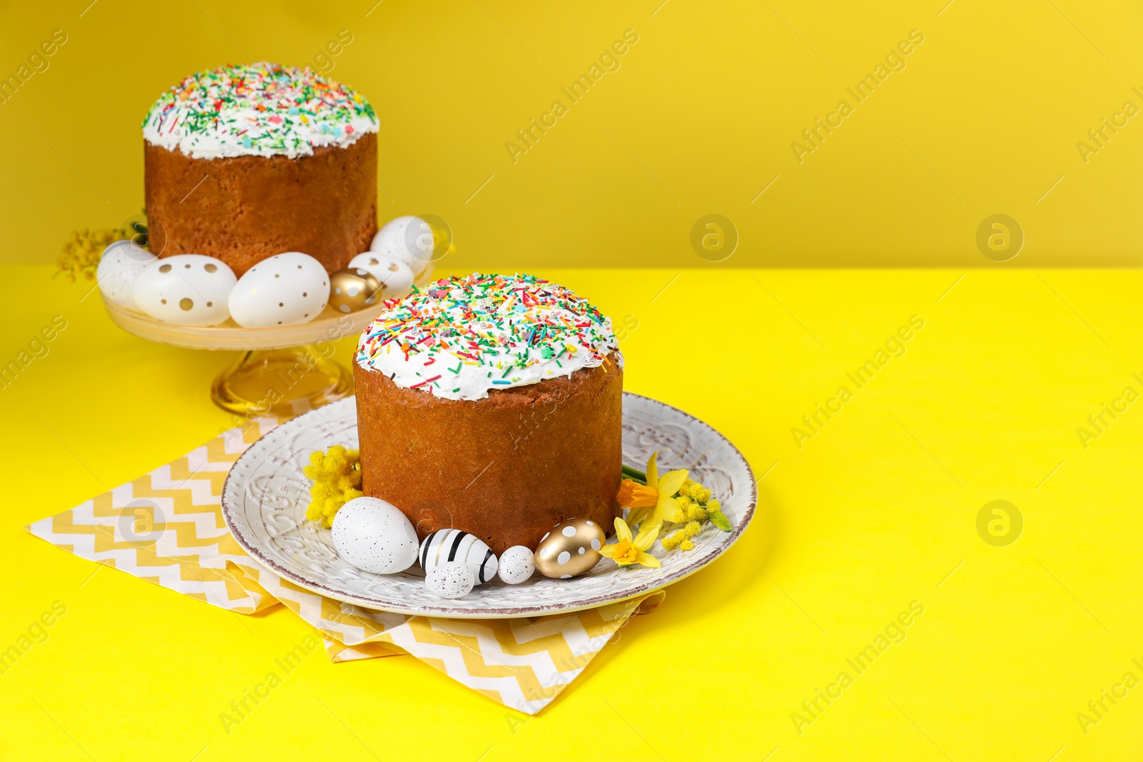 Photo of Traditional Easter cakes with sprinkles, painted eggs and beautiful spring flowers on yellow background, space for text