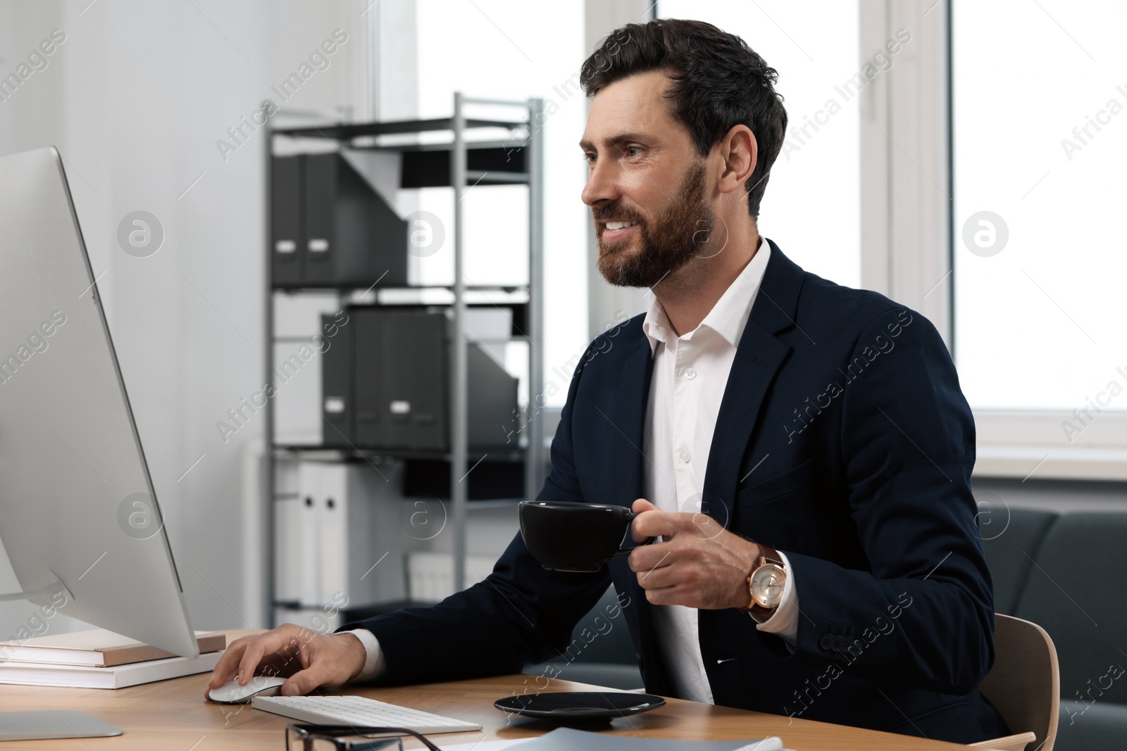 Photo of Handsome bearded man with cup of drink working on computer at table in office