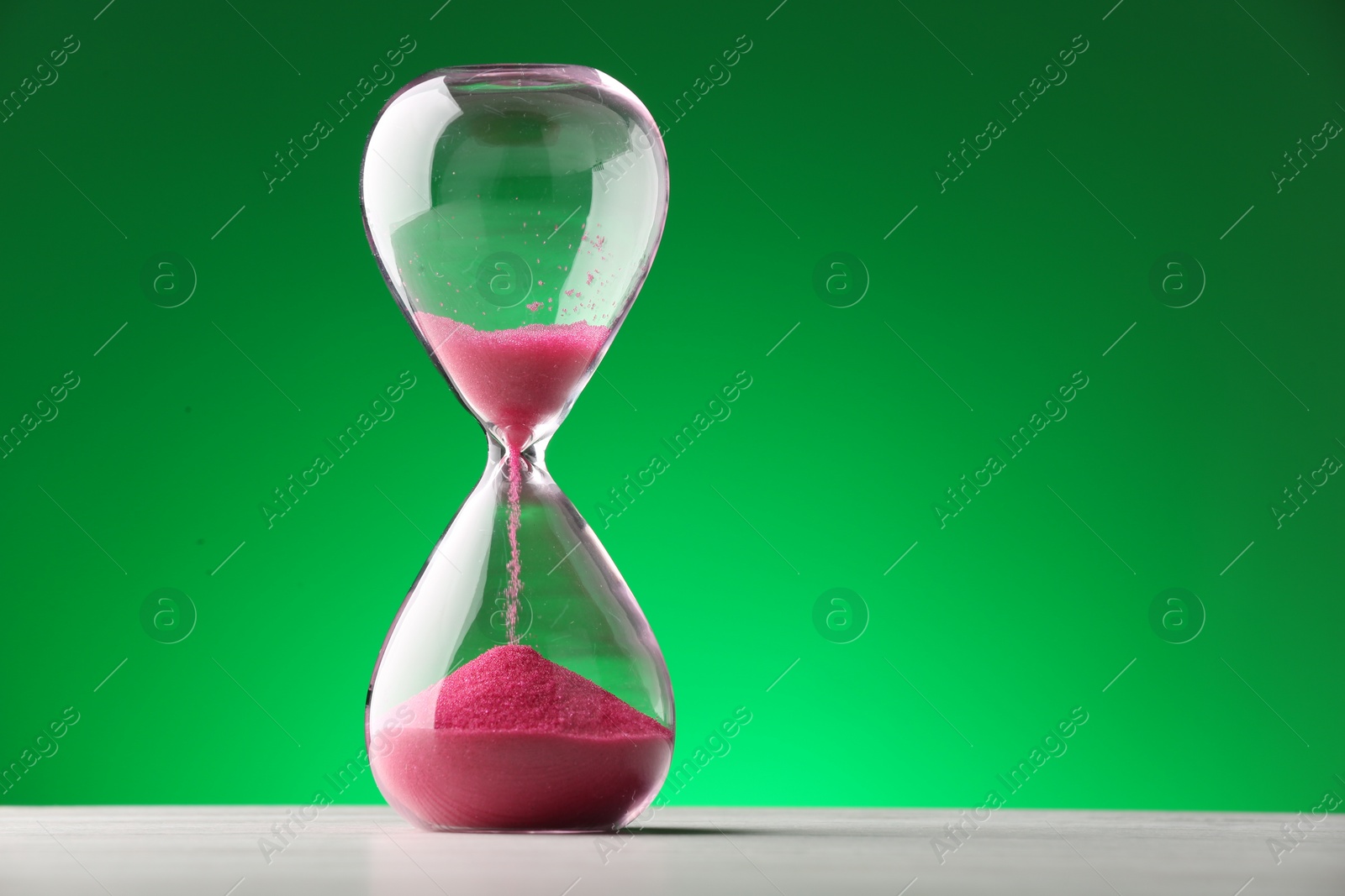 Photo of Hourglass with pink flowing sand on table against green background. Space for text