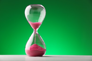 Hourglass with pink flowing sand on table against green background. Space for text
