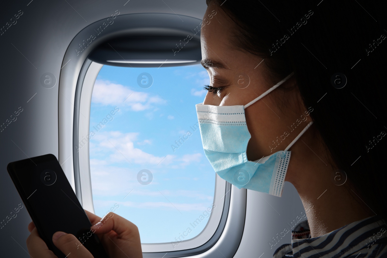 Image of Traveling by airplane during coronavirus pandemic. Woman with face mask and phone near porthole