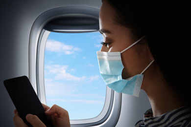 Traveling by airplane during coronavirus pandemic. Woman with face mask and phone near porthole