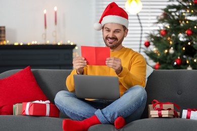 Celebrating Christmas online with exchanged by mail presents. Man in Santa hat reading greeting card during video call on laptop at home