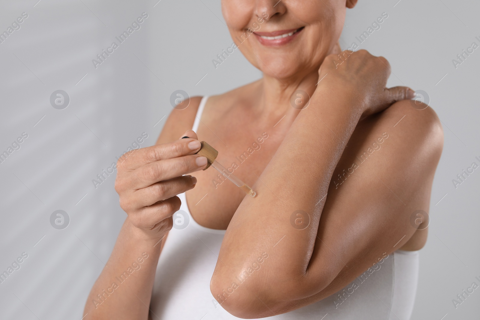 Photo of Happy woman applying body oil onto arm on grey background, closeup