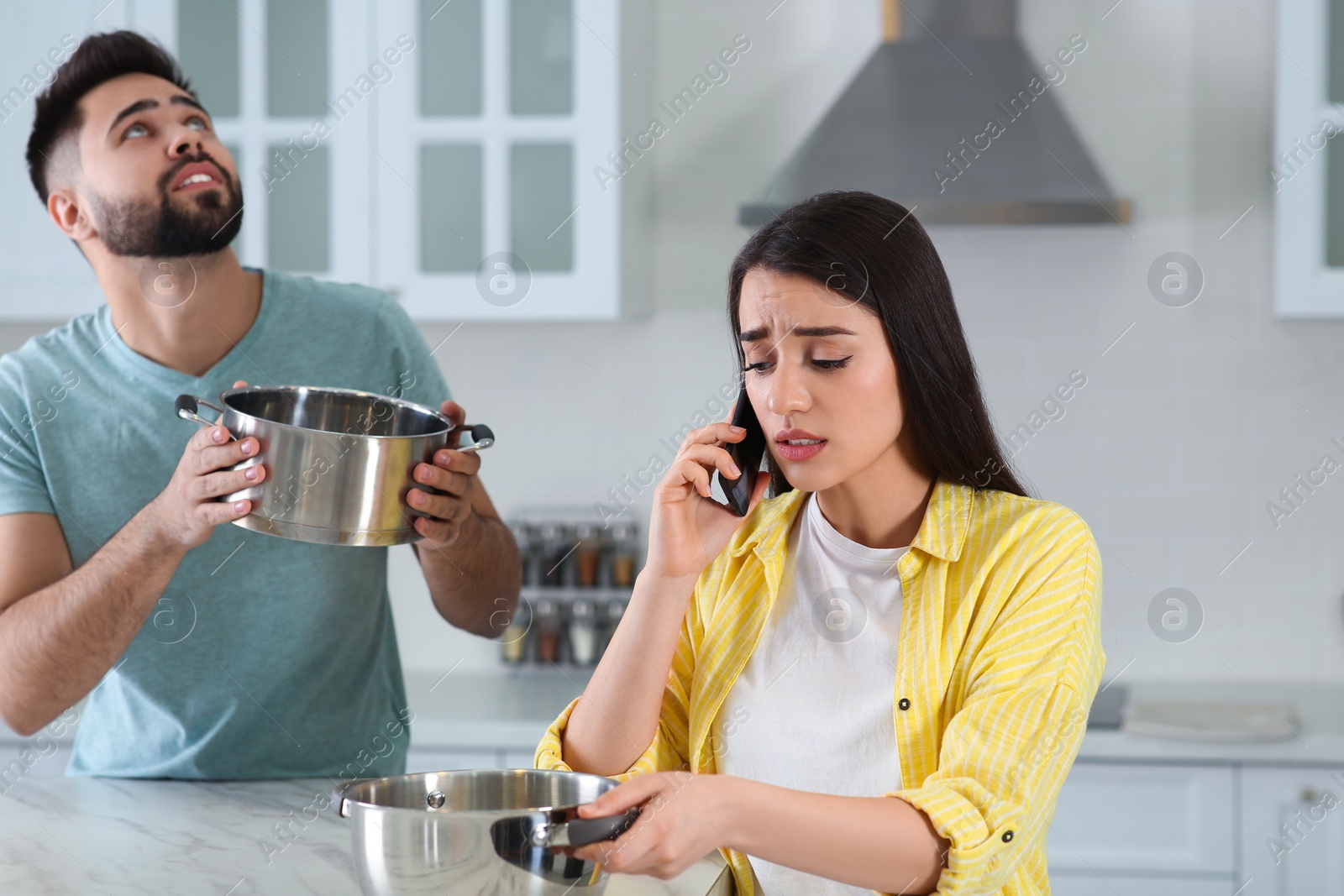 Photo of Young man collecting leaking water from ceiling while his girlfriend calling roof repair service in kitchen