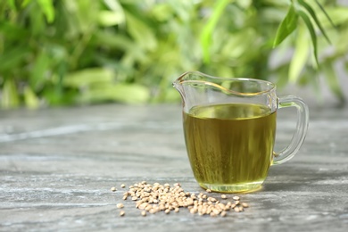 Glass pitcher with hemp oil on grey table against blurred background