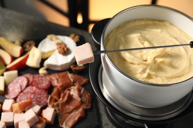 Photo of Fork with piece of ham, fondue pot with melted cheese and snacks on table, closeup