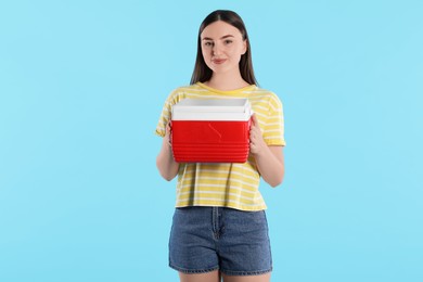 Beautiful young woman with plastic cool box on light blue background