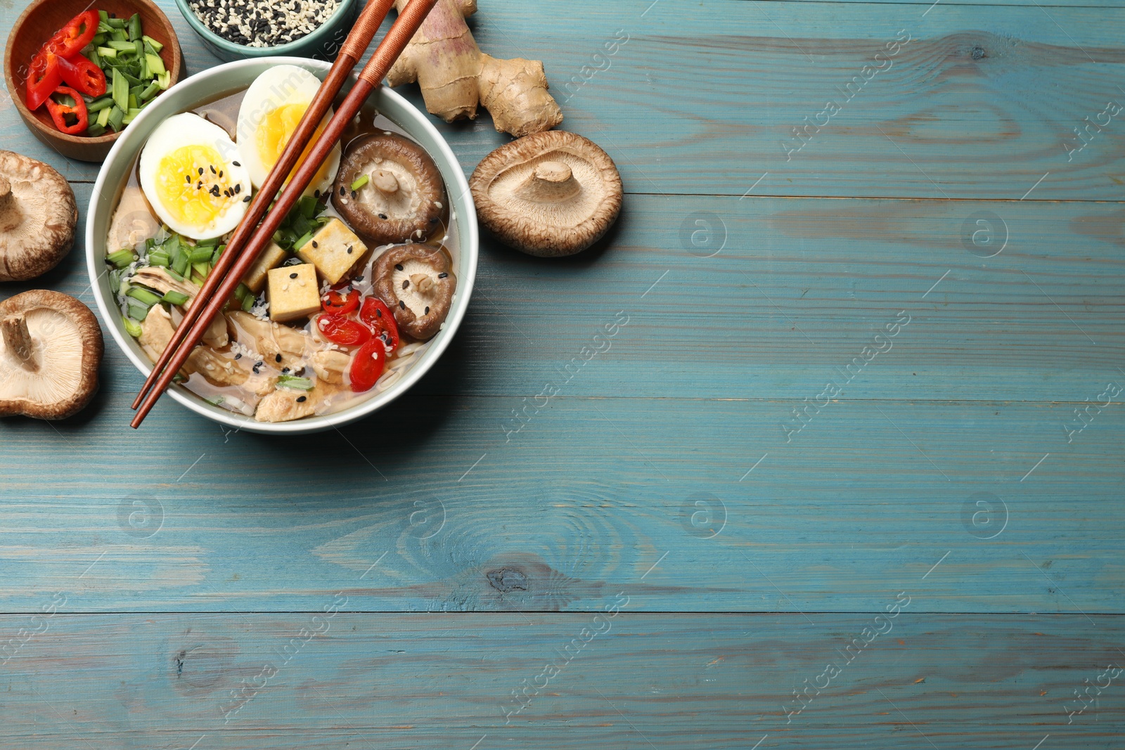 Photo of Bowl of delicious ramen and ingredients on light blue wooden table, flat lay with space for text. Noodle soup