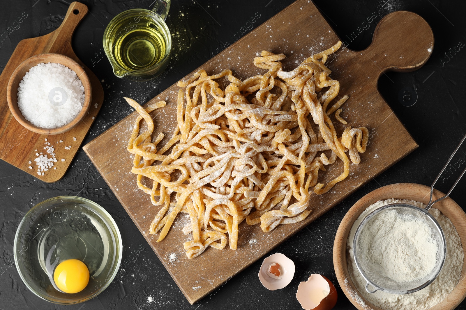 Photo of Board with homemade pasta, flour and ingredients on dark table, flat lay