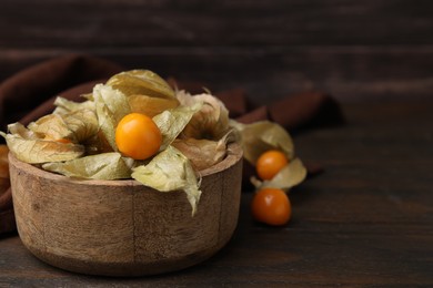 Ripe physalis fruits with calyxes in bowl on wooden table, space for text