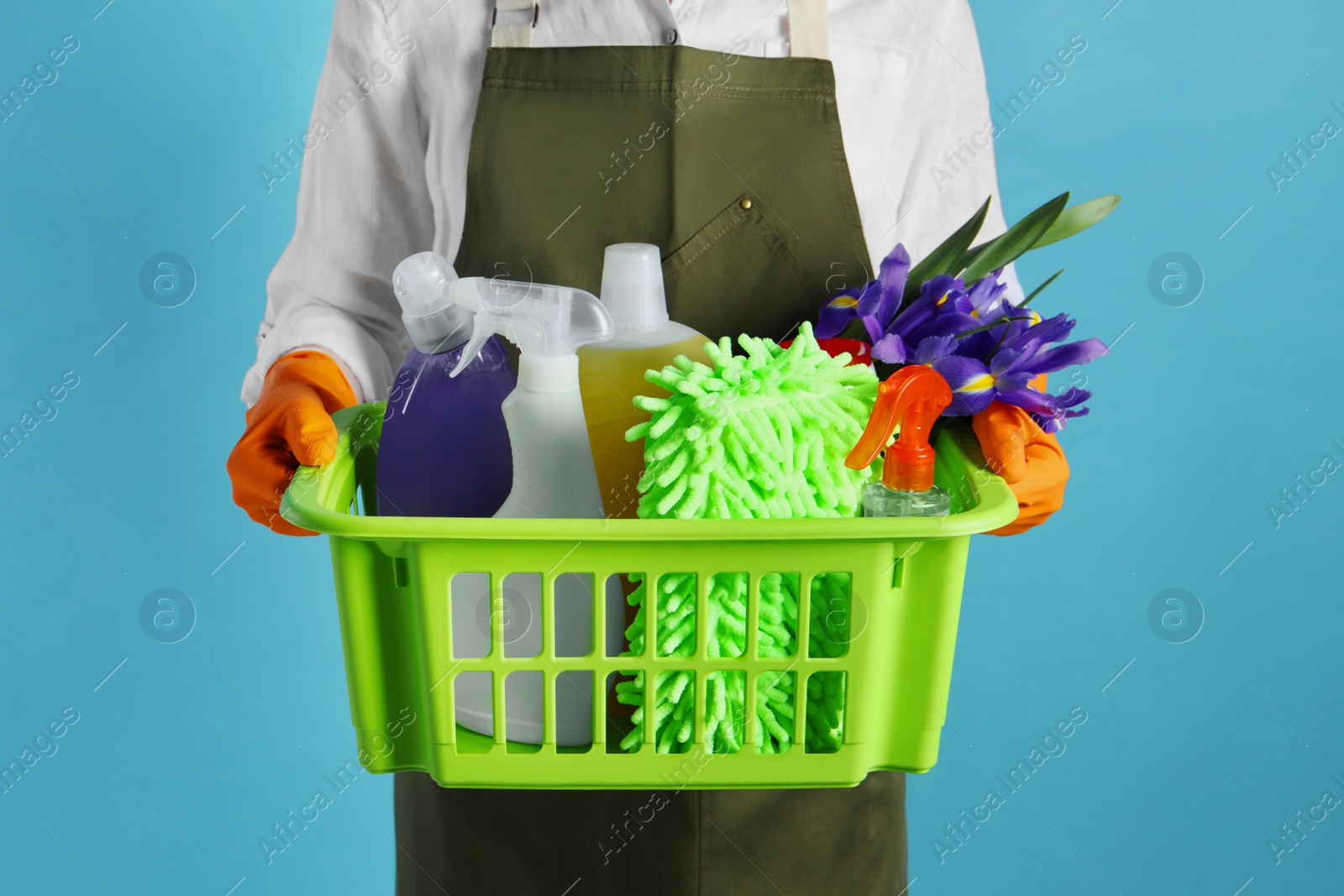 Photo of Spring cleaning. Woman holding basket with detergents, flowers and tools on light blue background, closeup