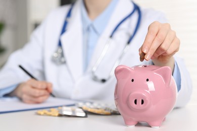 Photo of Doctor putting coin into piggy bank at white table indoors, closeup