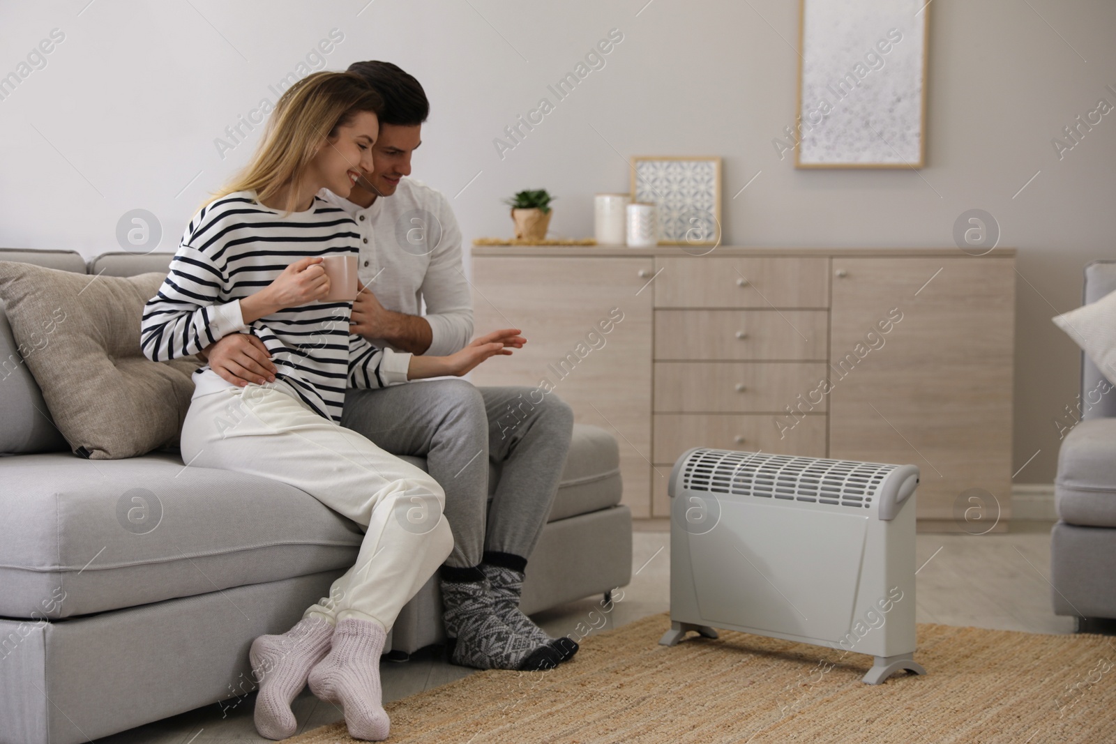 Photo of Happy couple sitting on sofa near electric heater at home