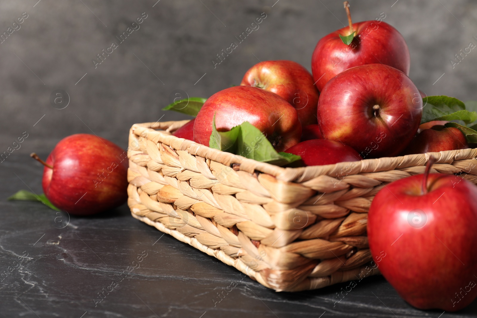 Photo of Fresh red apples and leaves in basket on dark grey table, closeup