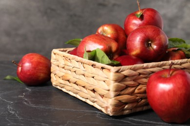 Photo of Fresh red apples and leaves in basket on dark grey table, closeup