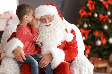 Photo of Little boy whispering in authentic Santa Claus' ear indoors