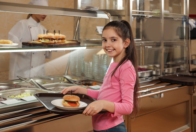 Photo of Little girl with plastic tray and burger near serving line in canteen. School food