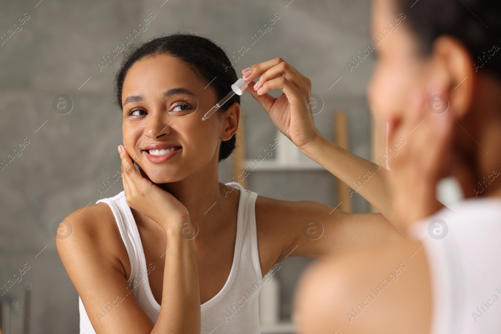 Photo of Smiling woman applying serum onto her face near mirror in bathroom