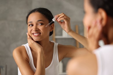 Photo of Smiling woman applying serum onto her face near mirror in bathroom