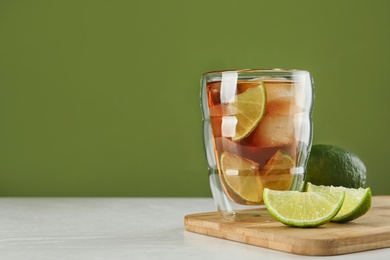 Glass of tasty ice tea with lime on white wooden table against green background, space for text