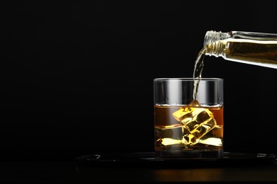 Pouring whiskey from bottle into glass with ice cubes at table against black background, space for text