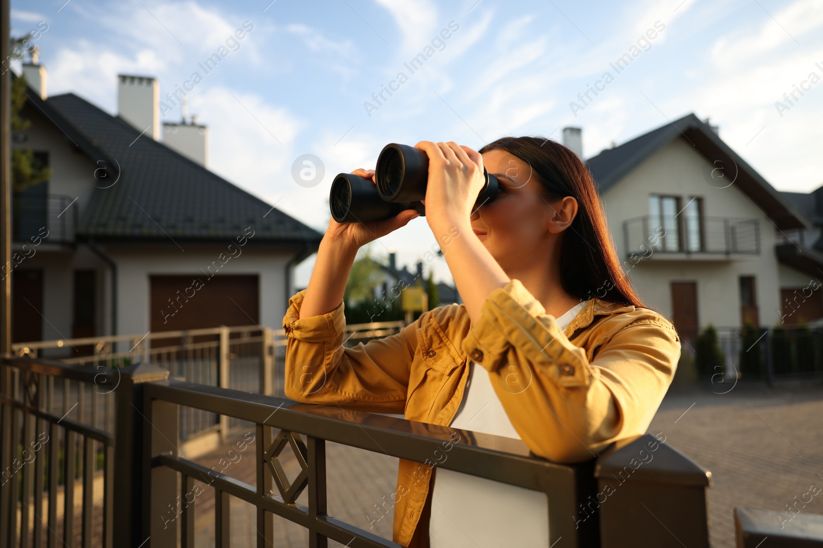 Photo of Concept of private life. Curious young woman with binoculars spying on neighbours over fence outdoors