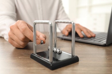 Photo of Man playing with Newton's cradle and using laptop at wooden table, closeup. Physics law of energy conservation