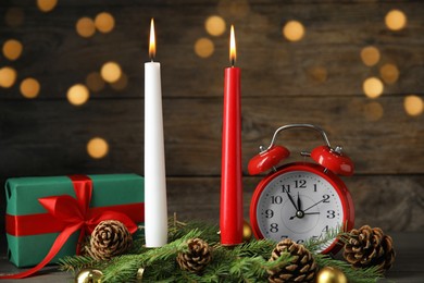 Photo of Burning candles, festive decor, gift box and alarm clock on wooden table against blurred Christmas lights