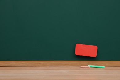 Photo of Colorful chalks on wooden table near green board with duster. Space for text